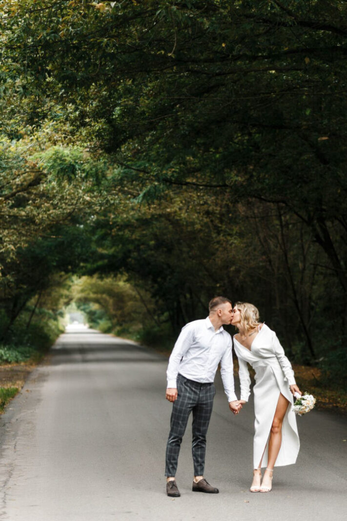 свадебная прогулка, жених и невеста целуются, держась за руку на форе арки из деревьев